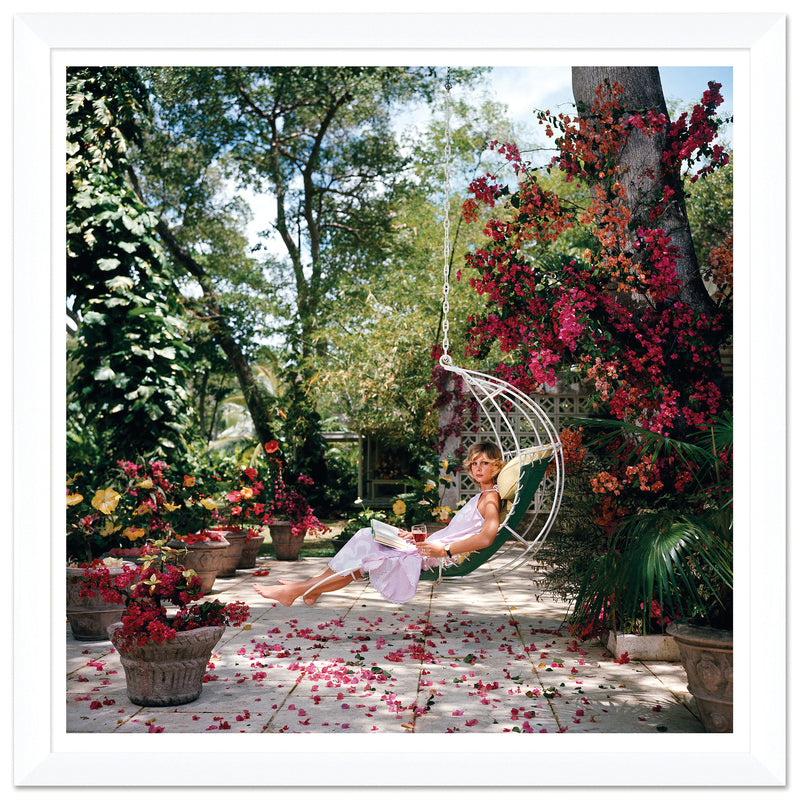 A person relaxes in a hanging chair in a garden, evoking the imagery of Soicher Marin's Slim Aarons By Getty Images Gallery "Ava Marshall," January 1, 1976, with vibrant flowers and potted plants adorning the stone patio.