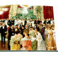 The open book "Legendary Costume Balls of the Twentieth Century" by Assouline reveals a painting of a formal ballroom, echoing Art Deco Paris, with elegantly dressed people conversing amid chandeliers and red drapery.