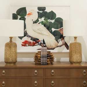 A dresser features two Visual Comfort Culloden Table Lamps in Marbled Sienna next to a stack of books. Above, vibrant parrot artwork adds color to the wall, perfectly balanced in height and width.