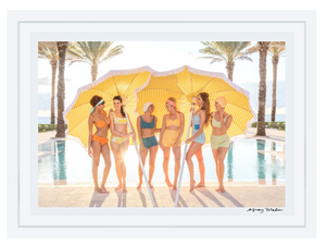 Six women in swimsuits stand by a pool under a large yellow-striped umbrella, echoing the Gray Malin Cabana Girls at The Breakers Palm Beach, with palm trees swaying gently in the background.