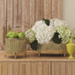 A wooden sideboard featuring two Small Brass Corrugated Bamboo Cachepots by Global Views—one with green apples, the other with white hydrangeas—stands beside a small yellow jar.