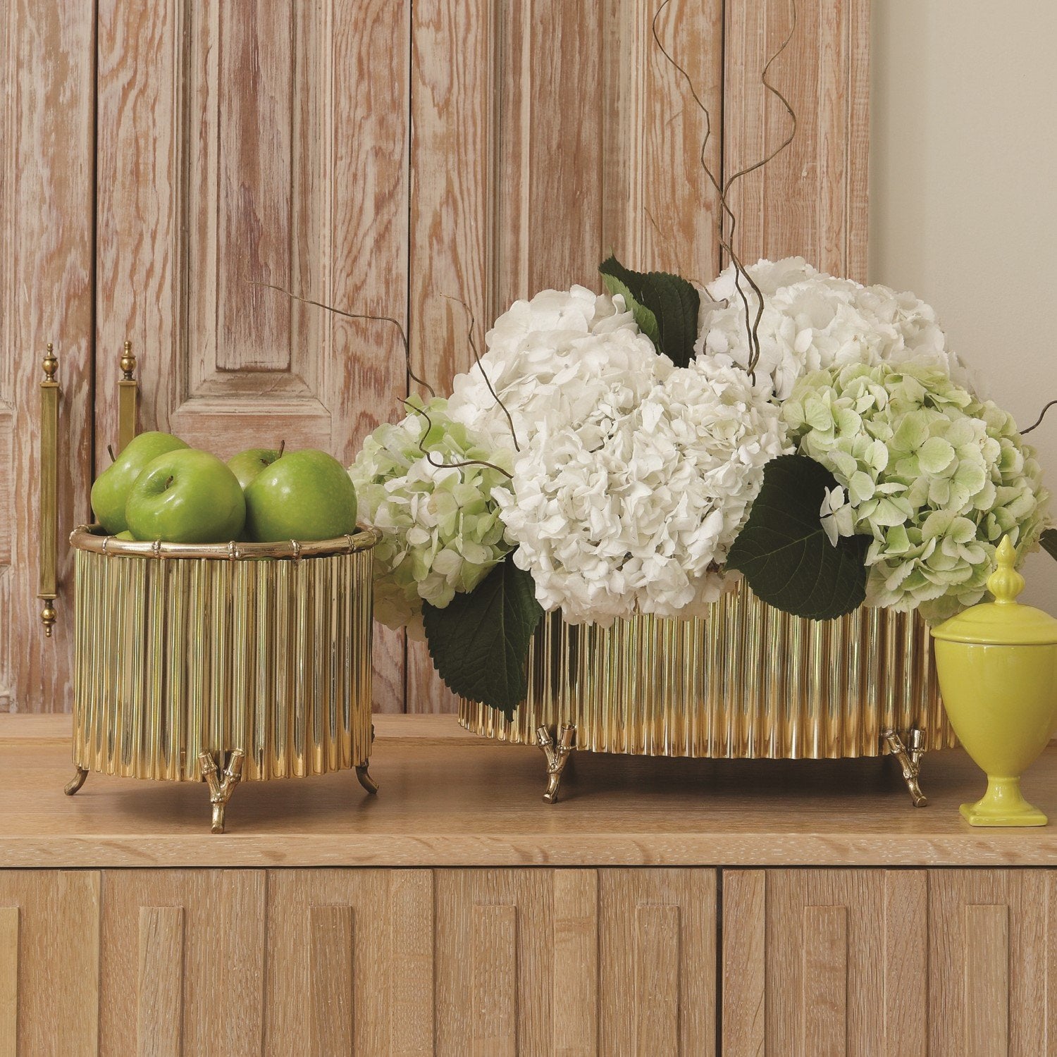 A wooden sideboard featuring two Small Brass Corrugated Bamboo Cachepots by Global Views—one with green apples, the other with white hydrangeas—stands beside a small yellow jar.