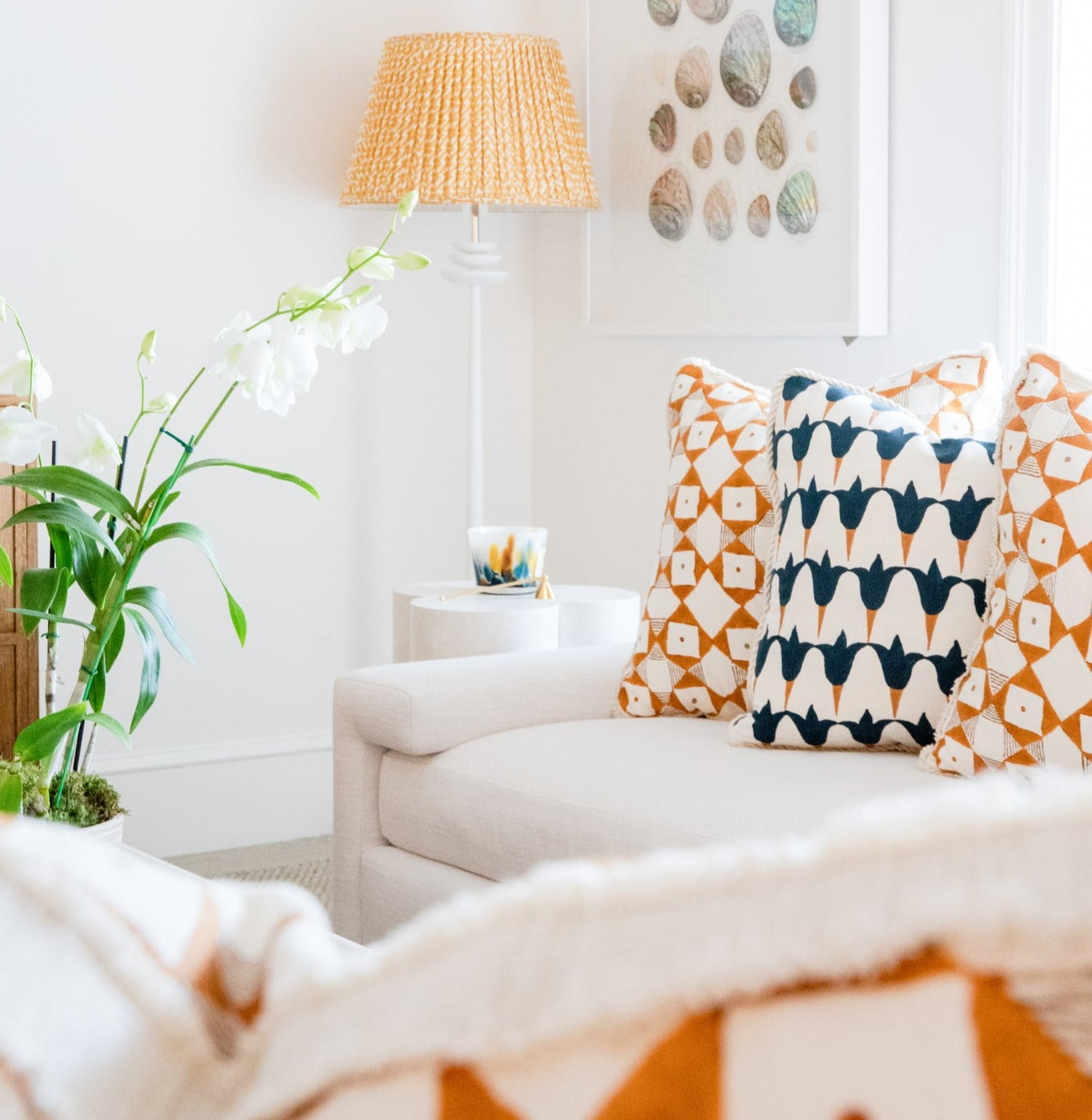 A cozy living room features a white sofa with 22" Hive Custom Pillows' Tulip Midnight & Copper pillows, a tall wicker lampshade, abstract wall art, and a potted orchid plant on a small table.