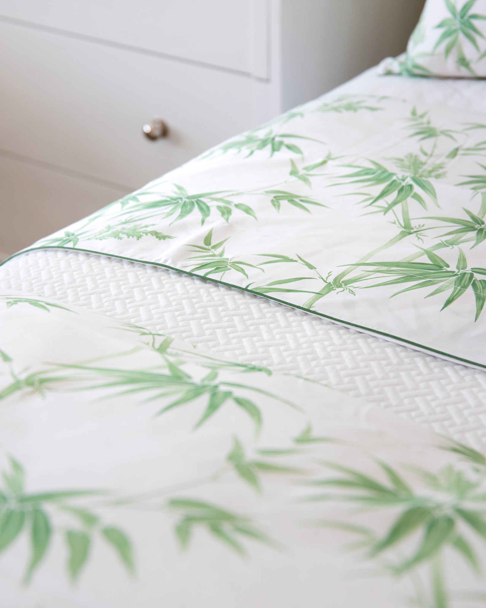 Close-up of a neatly made bed featuring Matouk’s Basketweave Bedding Collection in white, shown next to a white dresser with a silver knob.