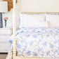 A neatly made bed with the Matouk San Cristobal Bedding Collection in Sky, white pillows, and a small decorative cushion; a white nightstand with a lamp, flowers, and a mirror is beside the bed.