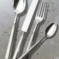 The Alain Saint Joanis Lignes 5 Piece Silver Plated Flatware Set, showcasing quality craftsmanship, is displayed on a light gray stone-textured surface.