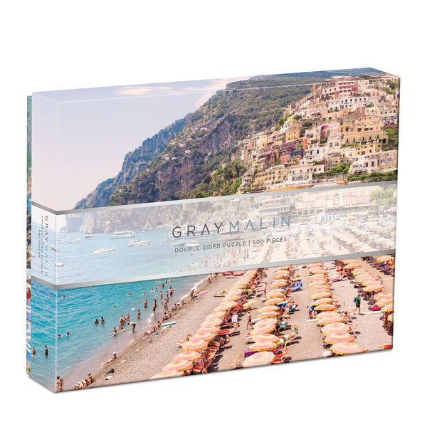 The Gray Malin The Italy Double-Sided Jigsaw Puzzle by Chronicle Books offers a coastal scene featuring vibrant beach umbrellas and hillside buildings, encapsulating the allure of an Italian coastal town.