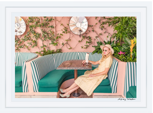 A woman in a vintage-style yellow dress and hat sits at a booth with green cushions, amidst greenery and mirrored wall decor, as featured in the Gray Malin "Girl at the Cabana Cafe" artwork, framed in white (29.5" x 41.5").