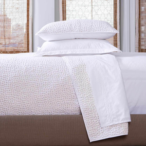 A neatly made bed features the John Robshaw Stitched Sand Coverlet, showcasing hand stitching, with two stacked pillows and white textured cotton voile bedding, set against windows framed by wooden blinds.