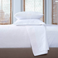 A bright room features a neatly made bed with John Robshaw's Stitched White Coverlet, crafted in India, paired with two stacked pillows and white cotton voile bedding, set against windows with woven blinds.