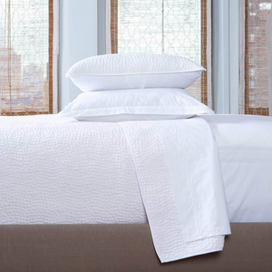 A bright room features a neatly made bed with John Robshaw's Stitched White Coverlet, crafted in India, paired with two stacked pillows and white cotton voile bedding, set against windows with woven blinds.