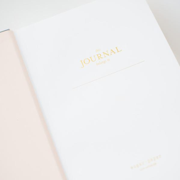 The Sugar Paper Tailored Chambray Journal is an open hardcover with a light pink cover, lined pages, gold "This journal belongs to" text on white paper.