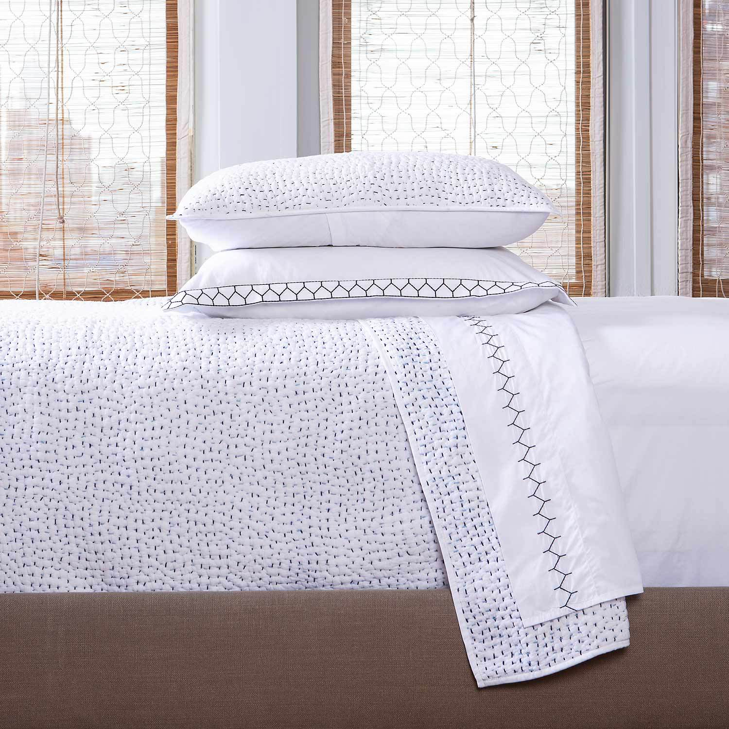 A neatly made bed with John Robshaw's Stitched Light Indigo Coverlet—a white cotton voile bedding with hand-stitched geometric patterns—plus two stacked pillows and large windows with bamboo blinds in the background. Made in India.