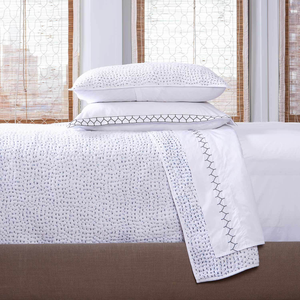 A neatly made bed with John Robshaw's Stitched Light Indigo Coverlet—a white cotton voile bedding with hand-stitched geometric patterns—plus two stacked pillows and large windows with bamboo blinds in the background. Made in India.