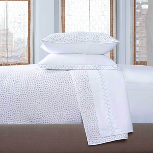 A neatly made bed with two stacked pillows features the Stitched Gray Coverlet by John Robshaw, hand-stitched with a subtle geometric pattern, set in front of three large windows with blinds.