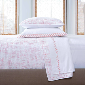 A neatly made bed with the John Robshaw Stitched Lotus Coverlet—white cotton voile featuring a subtle red pattern and hand stitching—two stacked pillows, and a folded blanket at the foot, set before tall windows with filtered light.