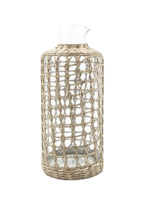 The Be Home Seagrass Cage Carafe features a clear cylindrical design with seagrass wrapping, a small spout, and an open top. Handwash is recommended to preserve its beauty.