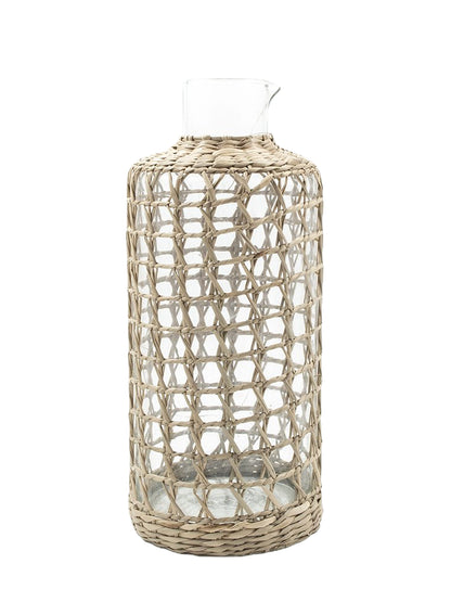 The Be Home Seagrass Cage Carafe features a clear cylindrical design with seagrass wrapping, a small spout, and an open top. Handwash is recommended to preserve its beauty.