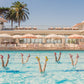 Eight people in a pool strike synchronized, high-heeled poses amid vibrant hotel scenes, palm trees, and umbrellas—captured in the Gray Malin Pool Party, White Frame (29.5" x 41.5") art piece by Gray Malin.