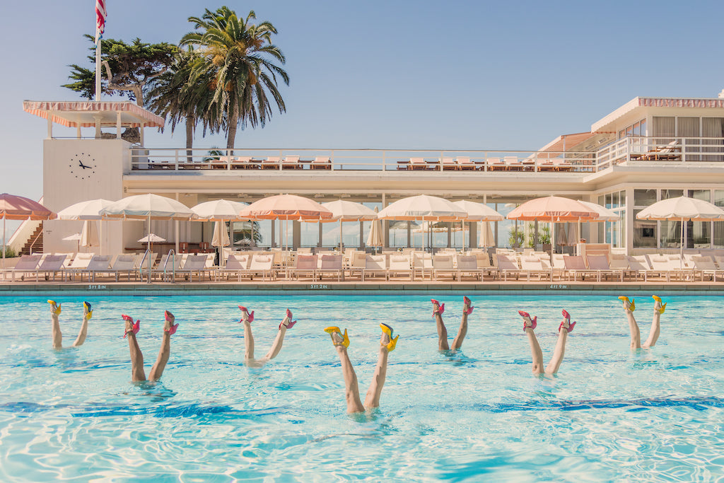 Eight people in a pool strike synchronized, high-heeled poses amid vibrant hotel scenes, palm trees, and umbrellas—captured in the Gray Malin Pool Party, White Frame (29.5" x 41.5") art piece by Gray Malin.