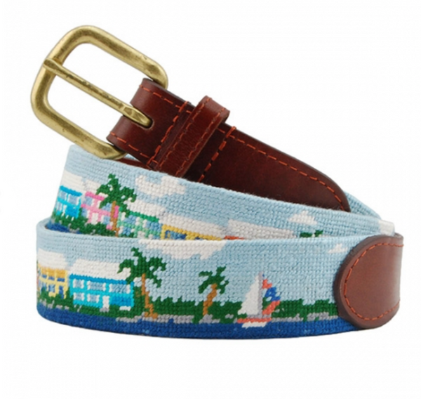 A Smathers & Branson Island Time Belt from Smathers & Branson, featuring a light blue needlepoint with a colorful seaside scene, including palm trees, houses, and a sailboat, accented with hand-stitched leather ends and a brass buckle.