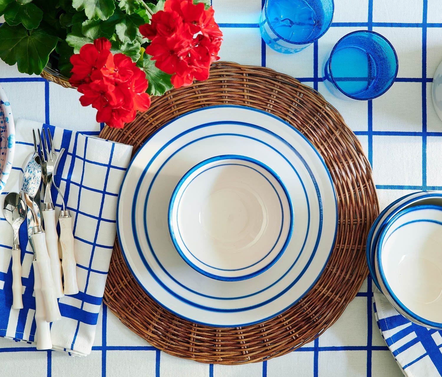 Table set with blue and white striped dishes, Blue Pheasant Montecito Acrylic Ivory 5 Piece Flatware Set, blue glasses, woven placemat, and red flowers on a blue grid-patterned tablecloth.