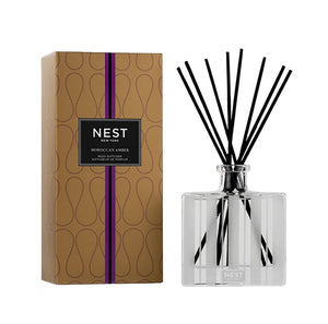 A Nest Moroccan Amber Diffuser set with a patterned box on the left and a clear glass bottle with black reeds on the right, featuring warm notes of amber and patchouli.
