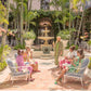 A group of six people in colorful attire gather around a fountain, capturing the essence of The Breakers' lush courtyard in Palm Beach, reminiscent of the Gray Malin "Palm Courtyard, The Breakers Palm Beach" artwork.