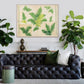 A black tufted leather sofa with green and white pillows sits before "Fern by Paule Marrot in Acrylic Box" by Natural Curiosities, flanked by potted plants and decorative vases on a white shelf.