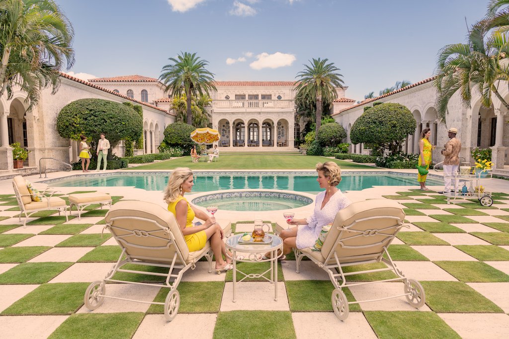 Two women relax by a pool in lounge chairs, sipping drinks amidst palm trees and a grand building reminiscent of Palm Beach style, creating a scene akin to "Gray Malin Poolside at La Follia, Palm Beach" by Gray Malin. Nearby, people walk and garden.