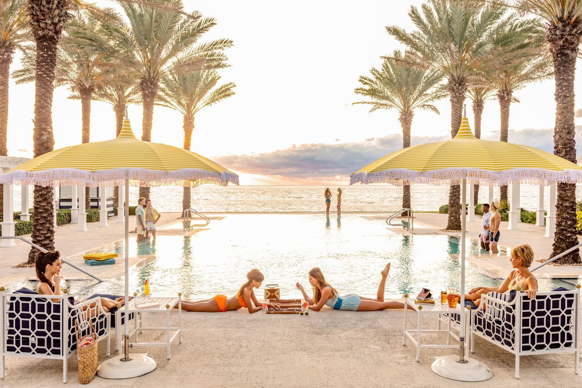 Relax by the pool under yellow umbrellas and palm trees with Gray Malin Poolside The Breakers Palm Beach, White Frame, 29.5" x 41.5", capturing an ocean view at sunset and a stylish scene of friends playing backgammon poolside.