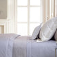 A neatly made bed with Yves Delorme’s Roma Blanc Fitted Sheet and multiple pillows sits next to a glass nightstand with a small lamp, set in a bright room with large windows.