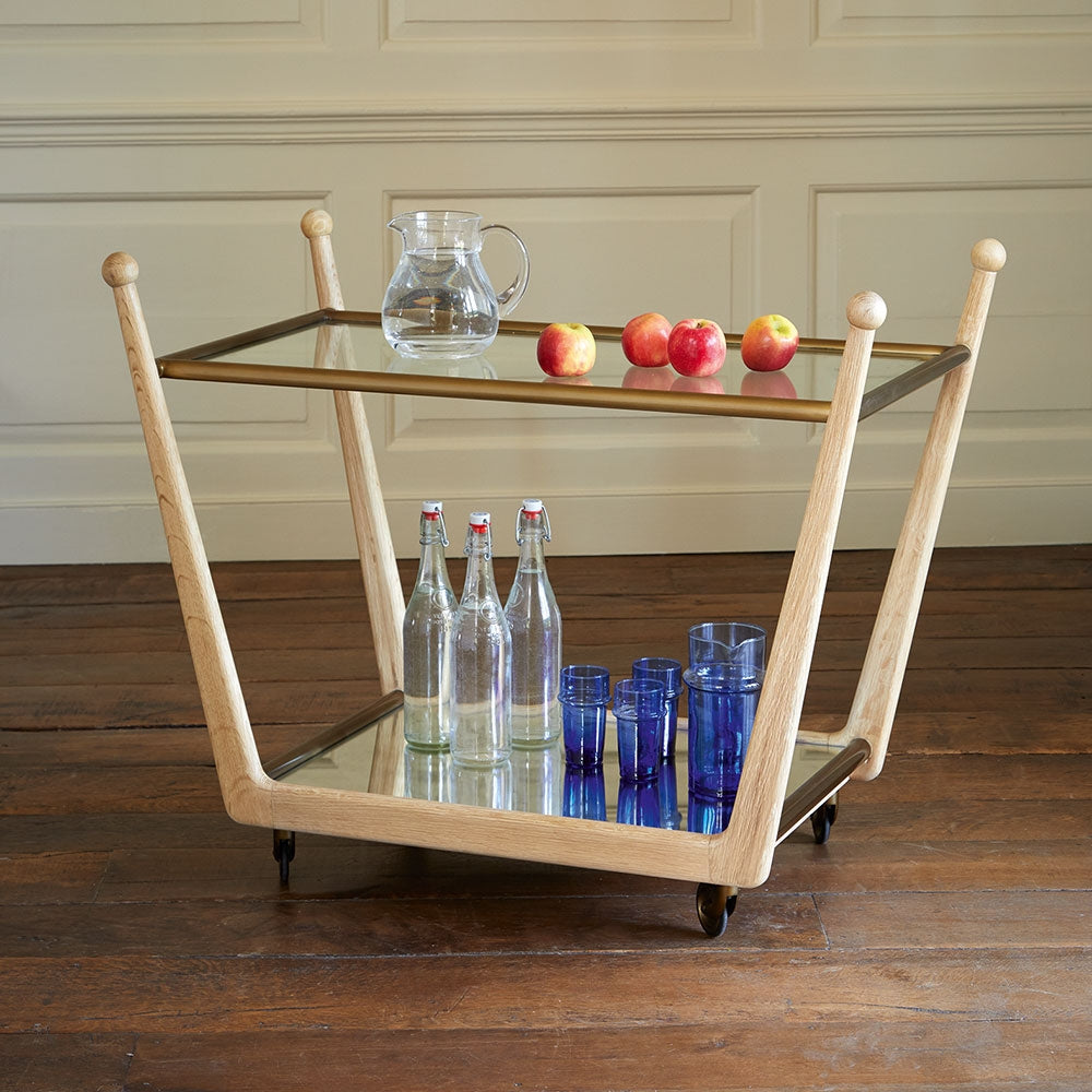A Rosa Trolley Cart in Bleached Oak from Julian Chichester boasts brass castors for easy mobility. It features two glass bottles and five azure blue high gloss vellum glasses on the bottom shelf, while a glass pitcher with four apples sits gracefully on the top shelf.