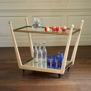 A Rosa Trolley Cart in Bleached Oak from Julian Chichester boasts brass castors for easy mobility. It features two glass bottles and five azure blue high gloss vellum glasses on the bottom shelf, while a glass pitcher with four apples sits gracefully on the top shelf.
