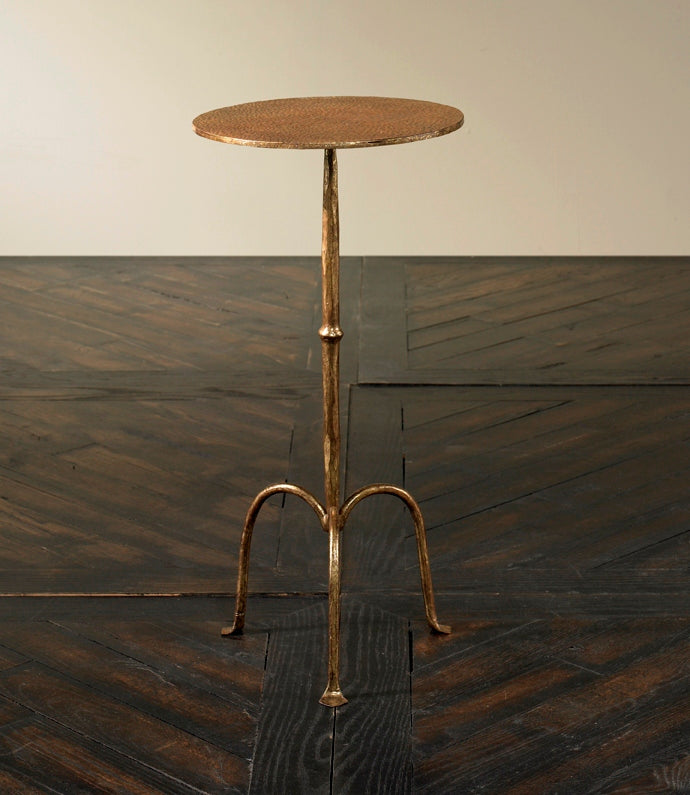 The Ruislip Side Table by Mr Brown stands elegantly on a dark, textured surface with its slender profile, round top, and three curved legs. Its Verona Gold finish shimmers subtly in the light.