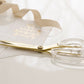 Elegant luxe Russell & Hazel gold & acrylic scissors beside a calligraphy-embellished wedding invitation and a satin ribbon.