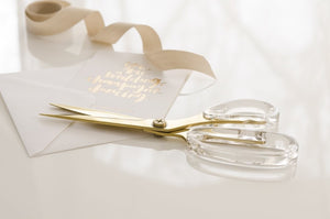 Elegant luxe Russell & Hazel gold & acrylic scissors beside a calligraphy-embellished wedding invitation and a satin ribbon.