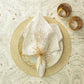 A decorative place setting features a gold and white plate, a cream napkin with the Kim Seybert Spider Bead Burst Champagne Napkin Ring, and two gold-rimmed glasses on a marbled surface.