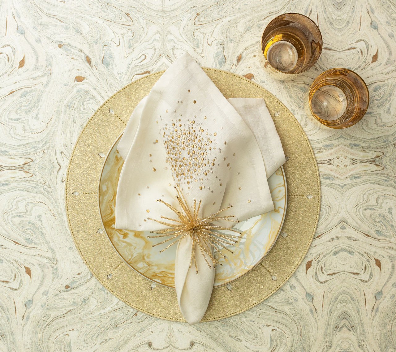 A decorative place setting features a gold and white plate, a cream napkin with the Kim Seybert Spider Bead Burst Champagne Napkin Ring, and two gold-rimmed glasses on a marbled surface.