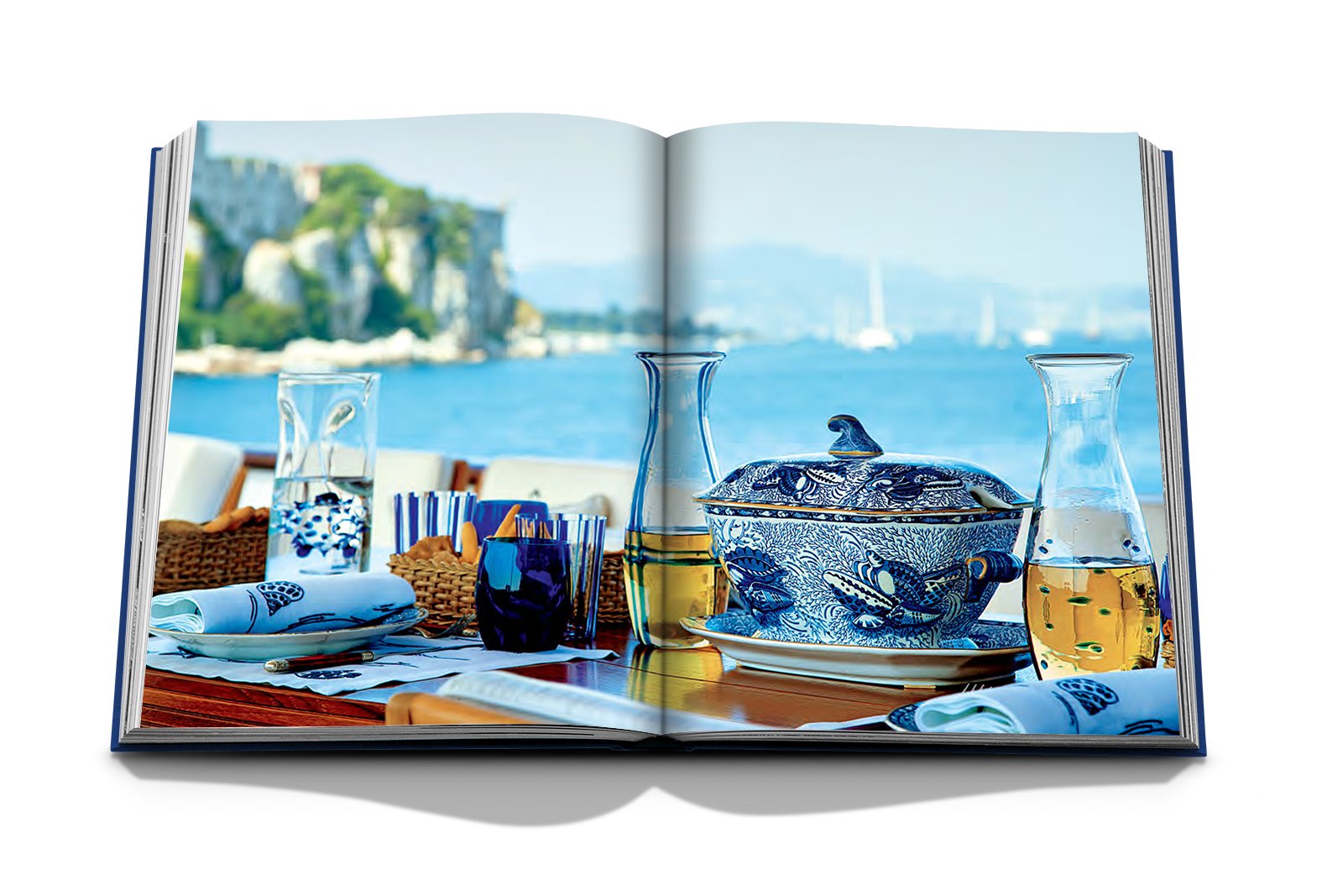 Valentino: At the Emperor's Table by Assouline showcases a seaside dining scene with blue-themed tableware, a patterned soup tureen, and glasses, all arranged as a haute couture table setting against a scenic coastal view.