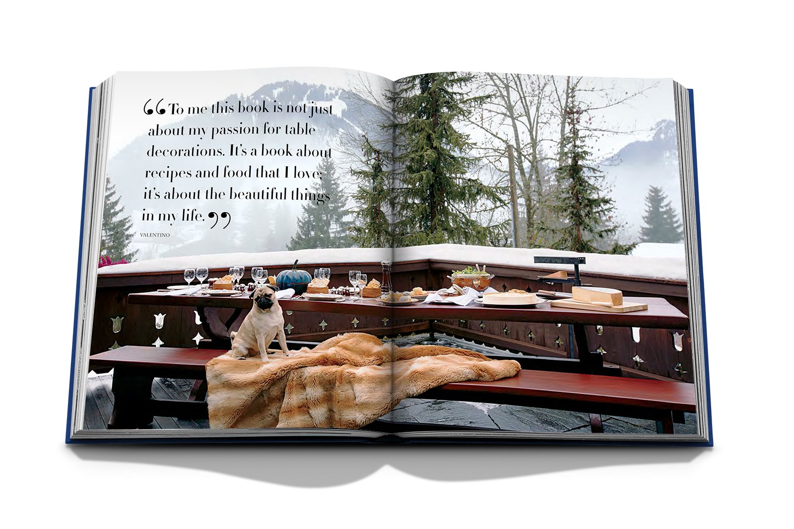 The Assouline book "Valentino: At the Emperor's Table" graces a balcony with haute couture table settings, food, drinks, and a cozy blanket. A small dog sits nearby, gazing at snow-covered trees in the background.
