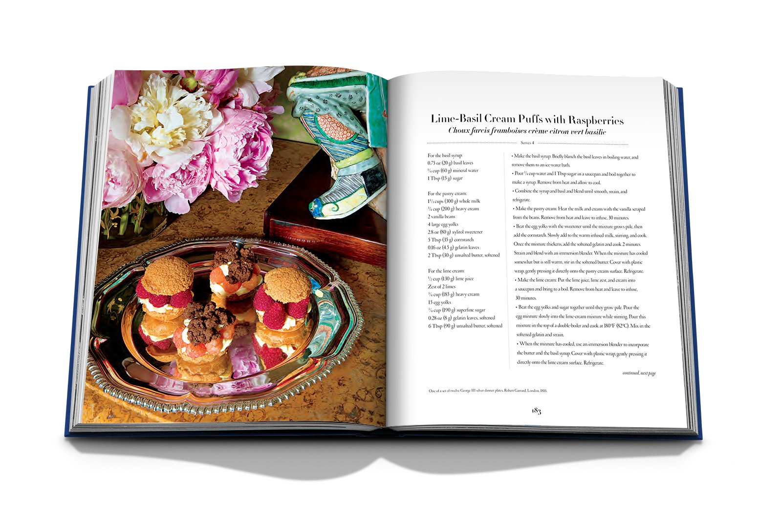 A copy of "Valentino: At the Emperor's Table" by Assouline is open to a lime-basil cream puffs with raspberries recipe, displayed next to an elegantly arranged dessert plate and a chic floral centerpiece, embodying haute couture aesthetics.