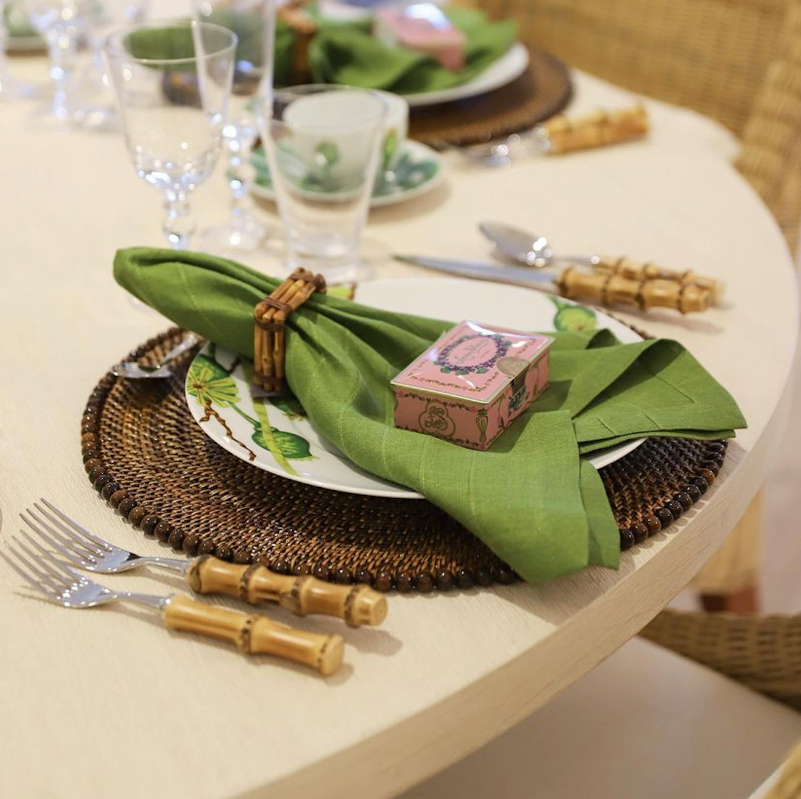 A place setting features Juliska’s Bamboo Napkin Ring with a green napkin, woven placemat, white plate, pink box, and bamboo-handled cutlery—a versatile accent adding authentic bamboo charm to your table.