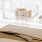 The Russell & Hazel Acrylic Tape Bloc, a clear acrylic and gold tape dispenser, rests atop a beige zippered case with a branded pen—a sleek addition to any desktop organizer.
