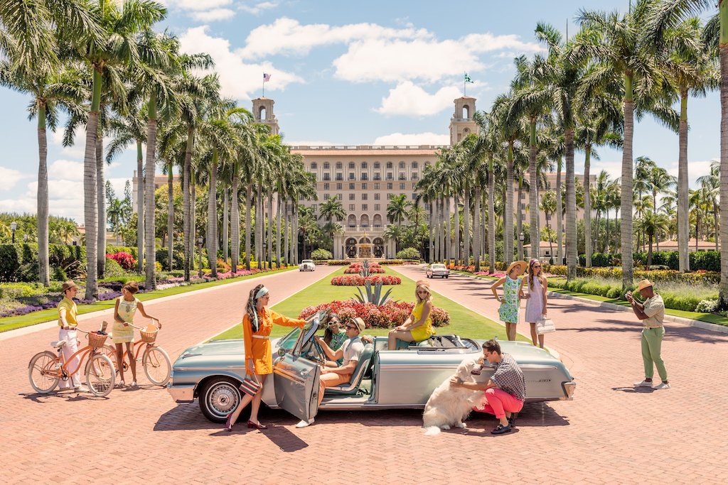 A group and a dog gather around a classic convertible on a palm-lined Palm Beach driveway, bikes leaning casually nearby, echoing the luxury lifestyle vibe seen in Gray Malin's "The Breakers Palm Beach," White Frame, 29.5" x 41.5".
