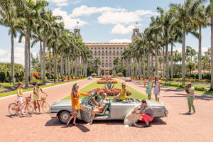 A group and a dog gather around a classic convertible on a palm-lined Palm Beach driveway, bikes leaning casually nearby, echoing the luxury lifestyle vibe seen in Gray Malin's "The Breakers Palm Beach," White Frame, 29.5" x 41.5".