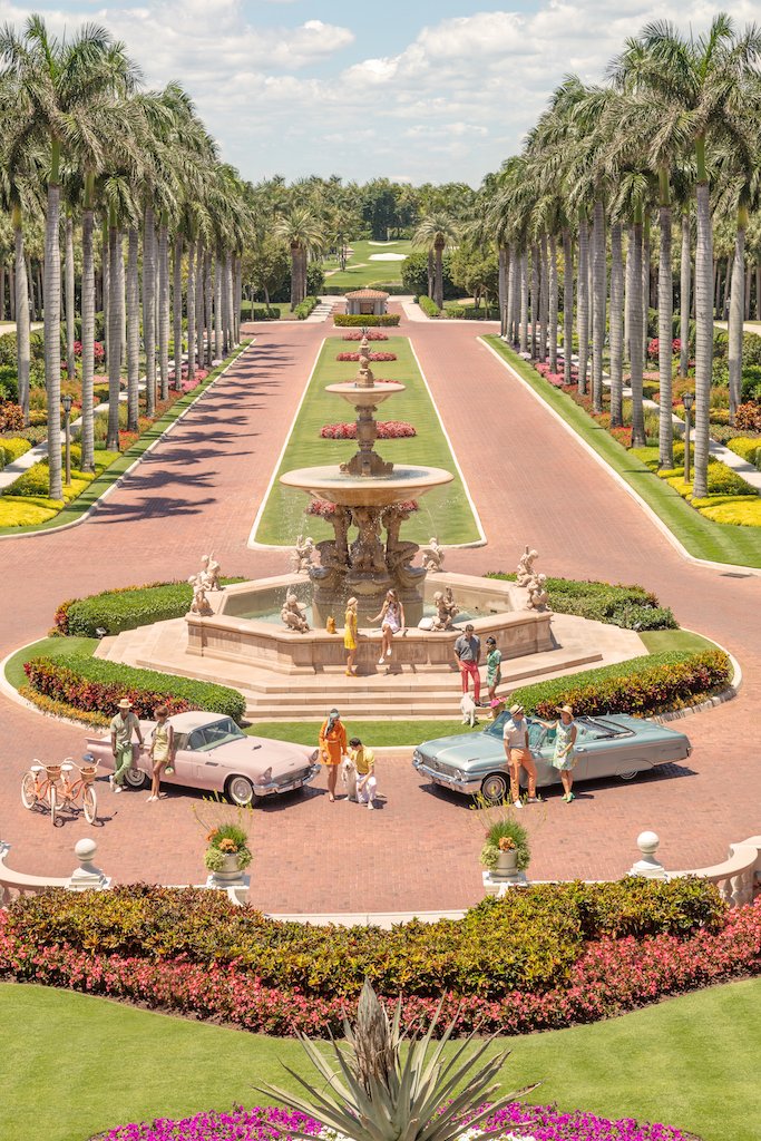 A group gathers by vintage cars and a fountain on the palm-lined main drive in "Gray Malin The Main Drive, The Breakers Palm Beach, White Frame, 24" x 36" by Gray Malin," with manicured gardens and a clear sky.