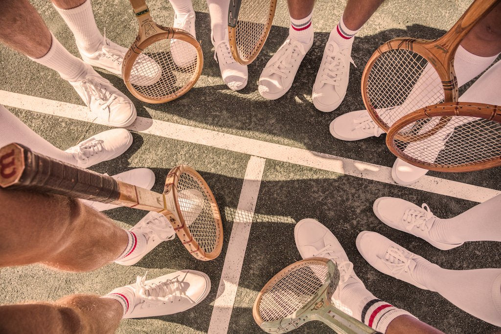 A tennis team in white shorts and shoes stands in a circle on the court, holding wooden rackets—evoking Gray Malin's "The Tennis Team Coral Beach Club" (White Frame, 29.5" x 41.5") from Gray Malin.