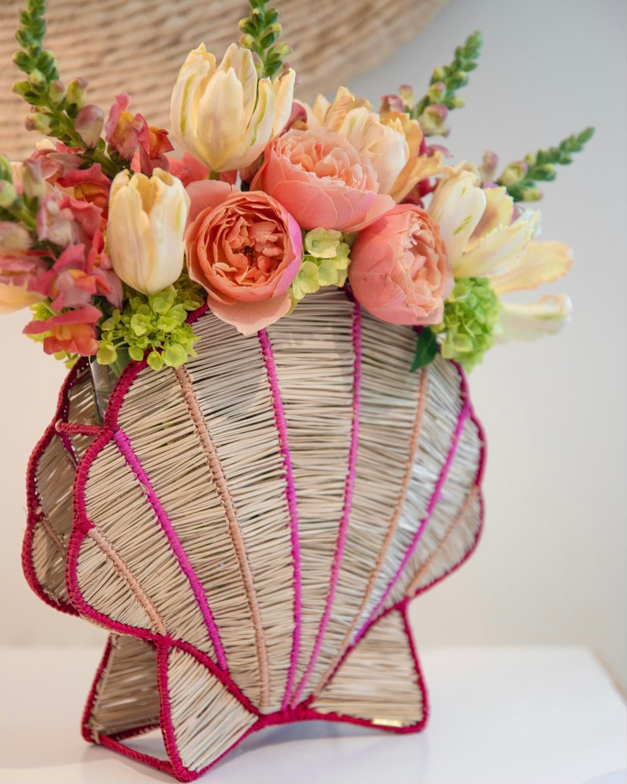 The Shell Flower Vase by Mercedes Salazar, crafted by skilled Bogotá artisans, holds a beautiful arrangement of pink and cream flowers with greenery on a white surface.