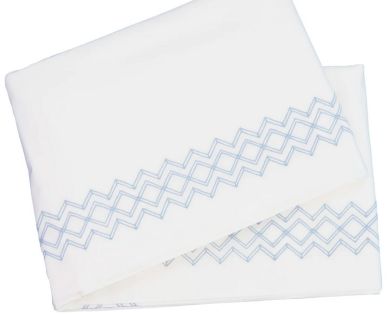 A pair of luxurious white bedsheets from the HIVE Exclusive Arezzo Bedding Collection by Matouk features cloud blue zigzag patterns along the edges, crafted from Egyptian cotton.